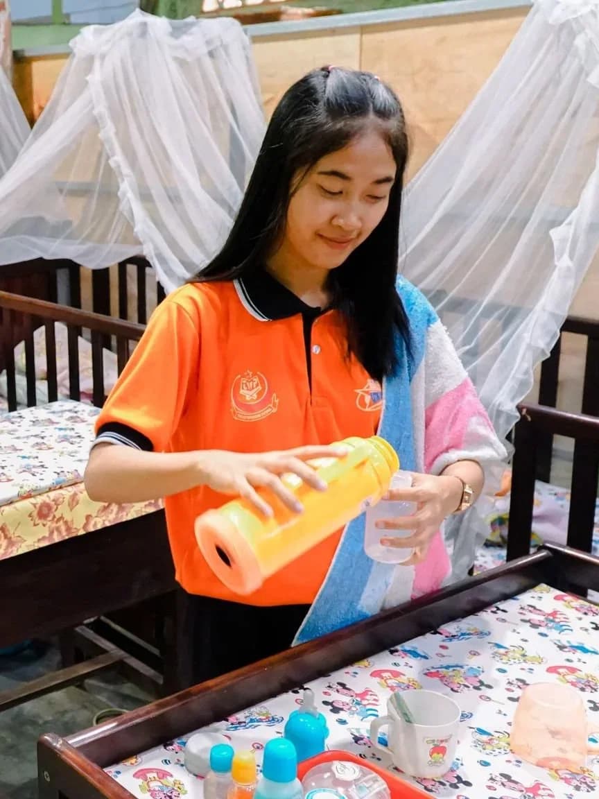 Trainee helpers practising newborn bottle feeding beside cots during infant care training at a training centre