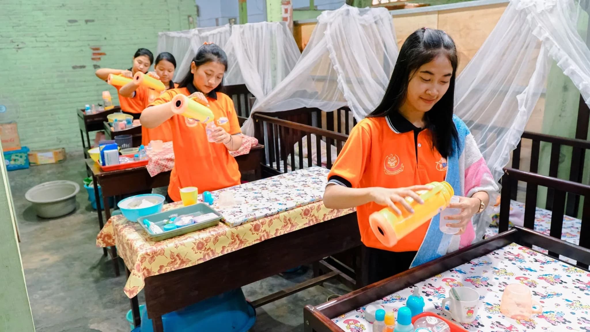 Trainee helpers practising newborn bottle feeding beside cots during infant care training at a training centre